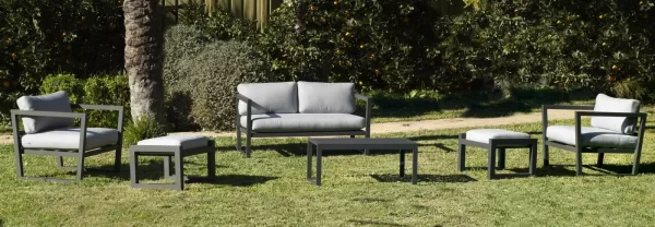 Mueble de exterior de jardín con un sofá de dos plazas ,dos sillones y una mesa de centro. El set de jardín está pintado en color gris.La imagen tiene platas en el fondo.