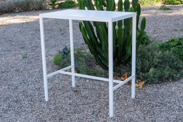 Mesa alta de exterior de 100 cm, modelo Caeli. Está fabricada en aluminio y pintada en color blanco. El tablero es de lamas de aluminio. La mesa se encuentra en un jardín rústico con suelo de piedras pequeñas y con arbustos al fondo.