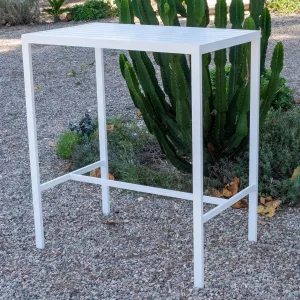 Mesa alta de exterior de 100 cm, modelo Caeli. Está fabricada en aluminio y pintada en color blanco. El tablero es de lamas de aluminio. La mesa se encuentra en un jardín rústico con suelo de piedras pequeñas y con arbustos al fondo.